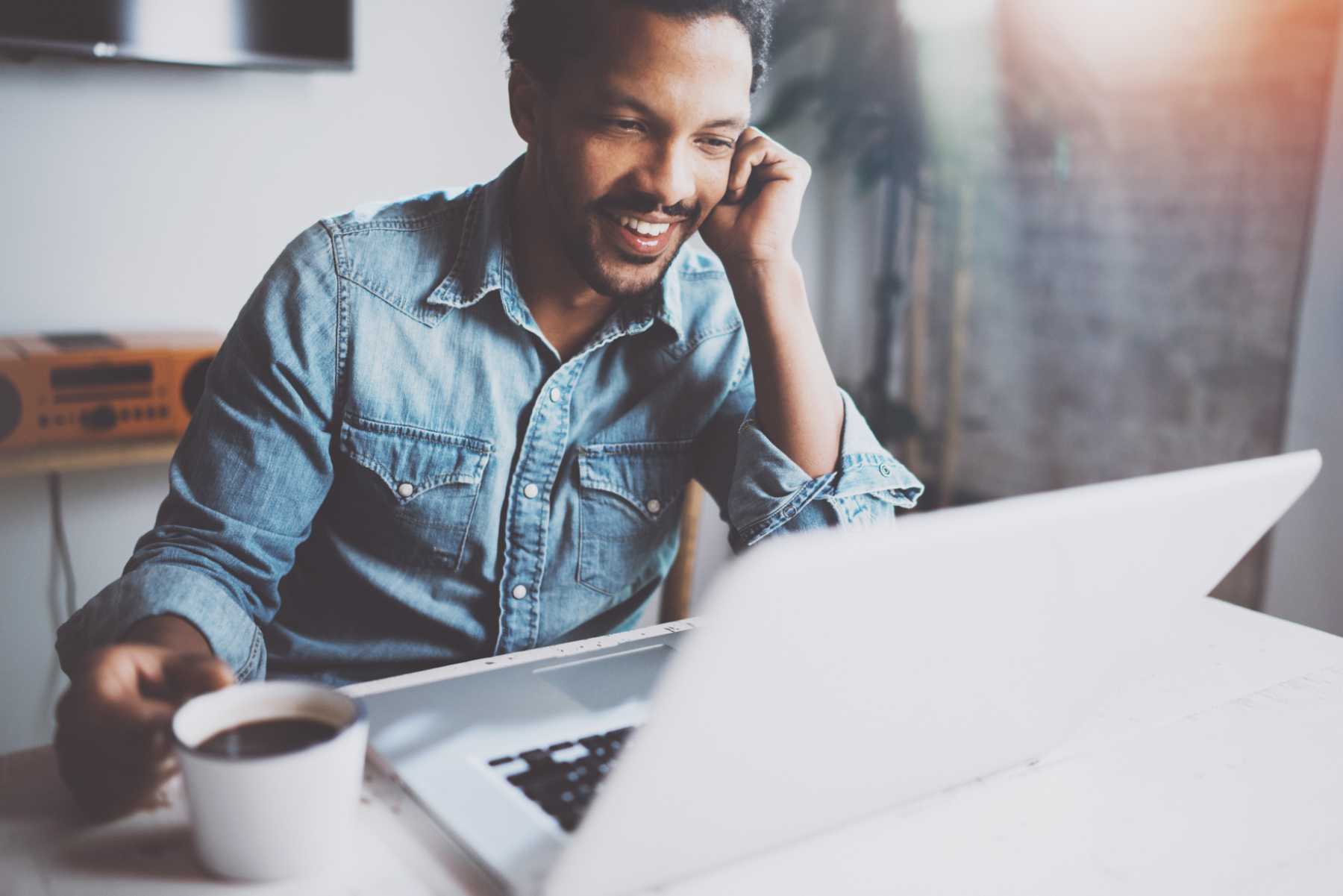 Happy bearded African man making video conversation via modern laptop with partners while holding white cup black coffee at home.Concept of young business people.Blurred background,flares effect